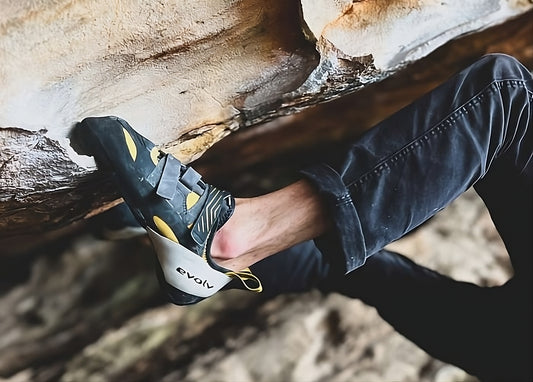 Close-up of a climber's foot wedged into a rock crevice, showcasing a black and yellow Evolv vegan climbing shoe with distinctive strap closures and the Evolv logo on the heel.