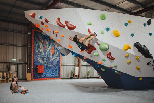 A climber at Portside Boulders Osborne Park is dynamically moving up a steeply angled bouldering wall, grasping onto brightly colored holds. Below, a spotter watches attentively, seated on the gym floor, ensuring safety during the climb.