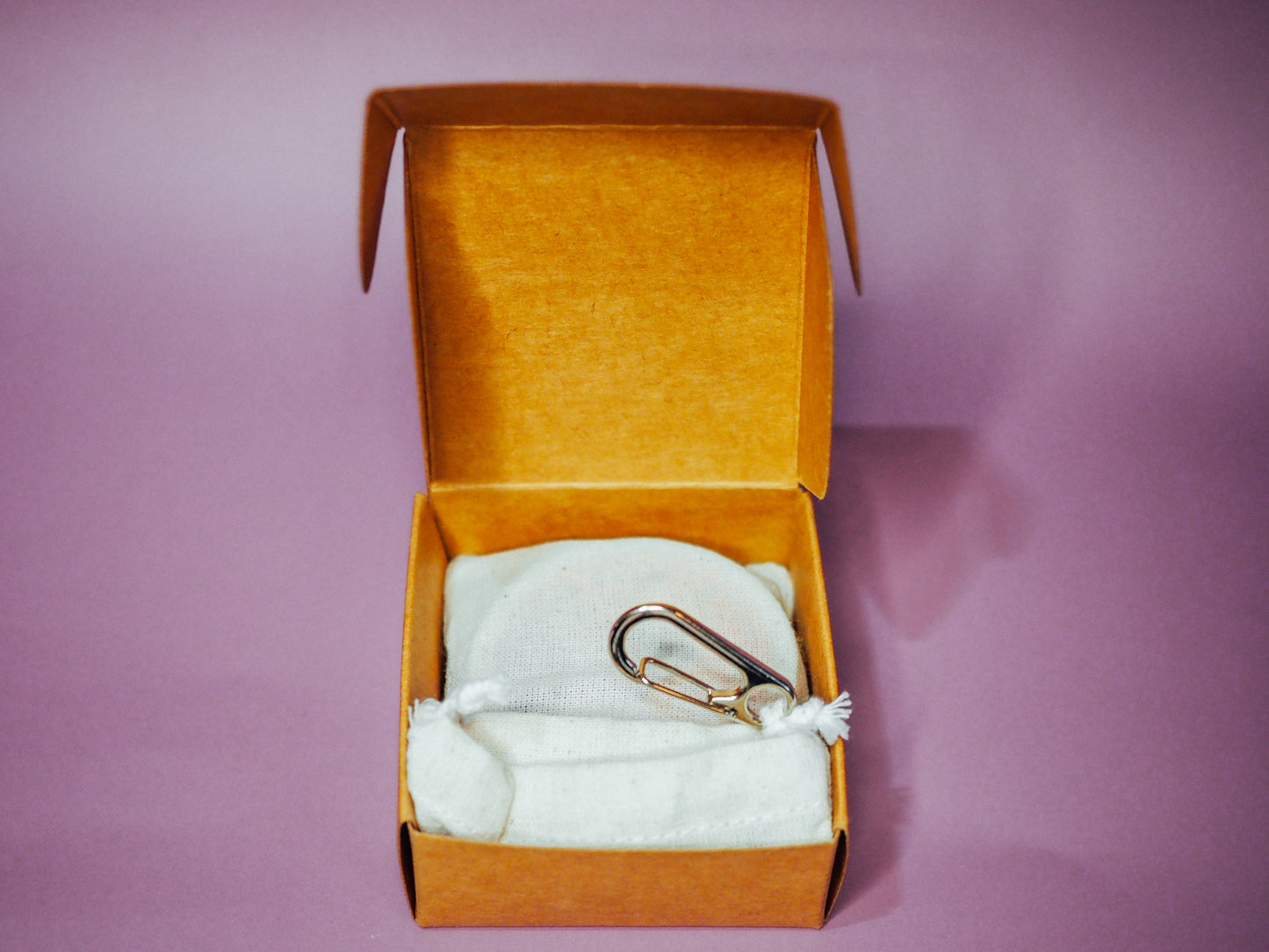 An overhead view of the open premium kraft box from Little Sammy's Climbers Balm Travel Bundle against a light purple backdrop. Inside, there's a neatly placed calico cotton bag with a silver carabiner, containing a 30ml climbers balm and bamboo spatula.