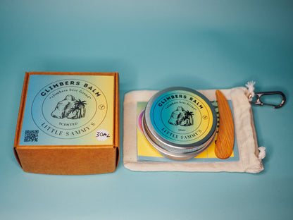 A scented travel bundle from Little Sammy's Climbers Balm, consisting of a 30ml tin displayed alongside a drawstring calico cotton bag with a wooden spatula and carabiner, set against a blue background. The focus is on the product contents and kraft box with vibrant labelling showcasing the product.