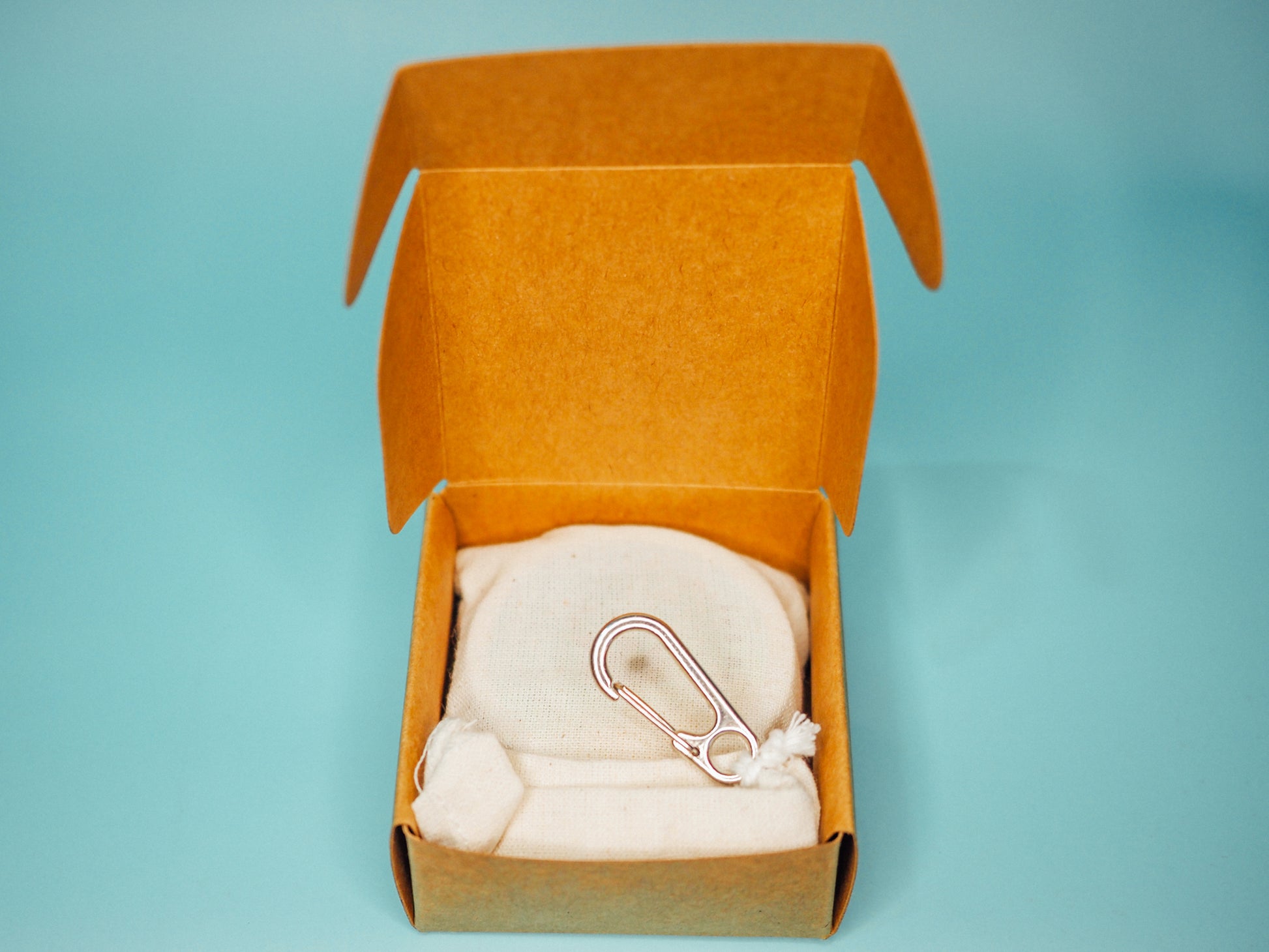 An overhead view of the open premium kraft box from Little Sammy's Climbers Balm Travel Bundle against a blue backdrop. Inside, there's a neatly placed calico cotton bag with a silver carabiner, containing a 30ml climbers balm and bamboo spatula.