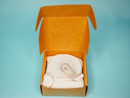 An overhead view of the open premium kraft box from Little Sammy's Climbers Balm Travel Bundle against a blue backdrop. Inside, there's a neatly placed calico cotton bag with a silver carabiner, containing a 30ml climbers balm and bamboo spatula.