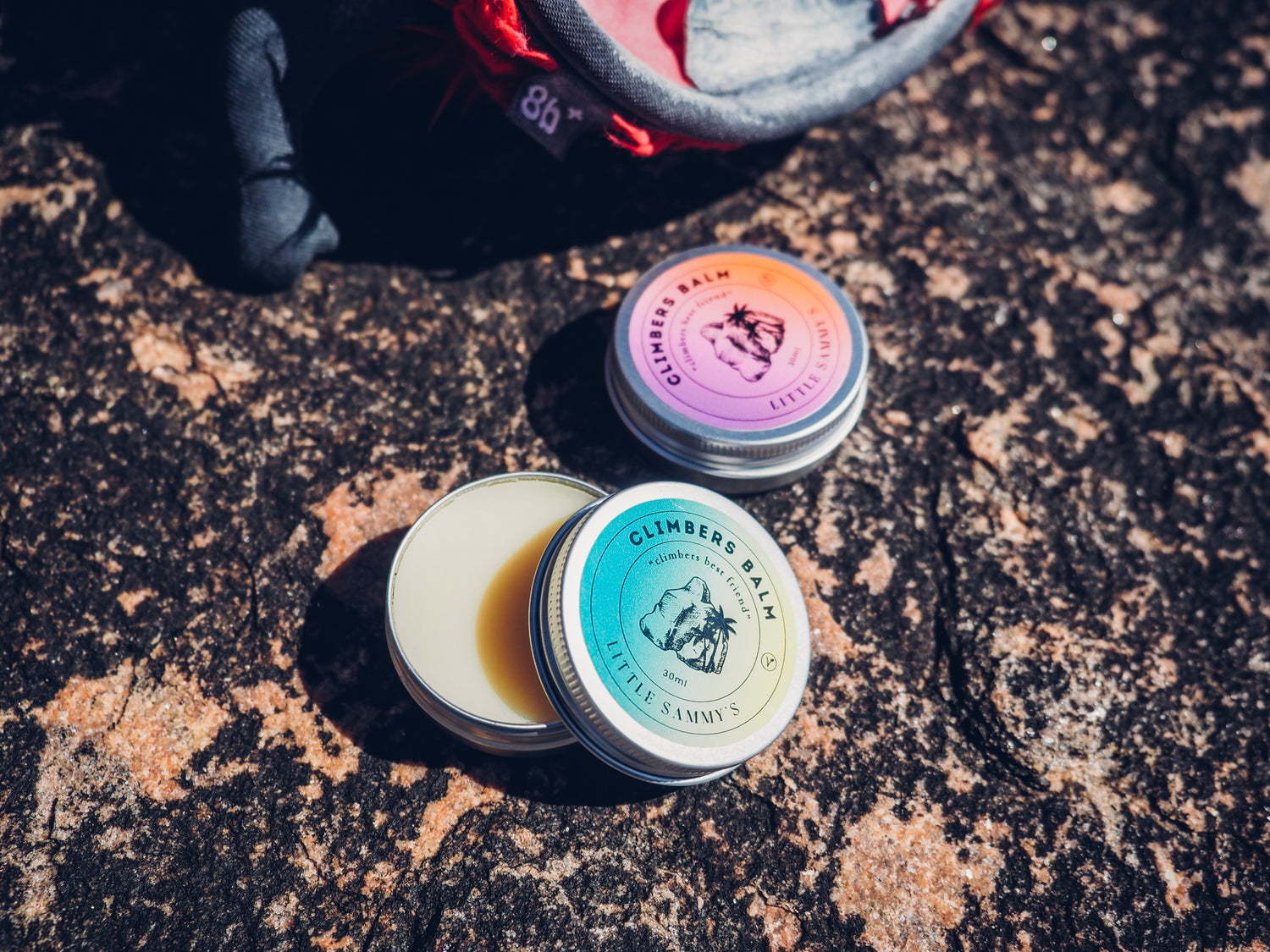 Close-up of Little Sammy's Climber's Balm tins (scented and unscented variants) resting on a textured rock surface at a climbing spot. The vibrant balm packaging contrasts with the rugged outdoor environment, showcasing an essential product for climbers' skincare and recovery.