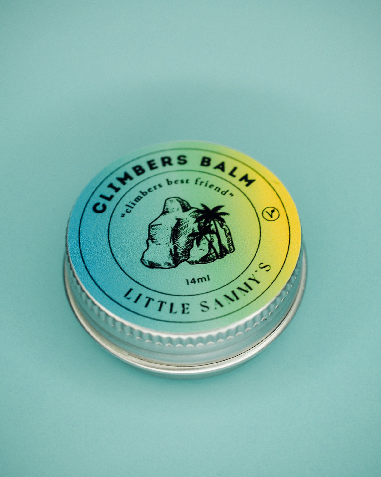 A 14ml tin of Little Sammy's Climbers Balm, closed and viewed from above, showcases the product's compact size. The top of the tin features a circular label with a gradient from blue to yellow, including an illustration of a boulder and a palm tree. The text reads 'CLIMBERS BALM - climbers best friend' with '14ml' and 'LITTLE SAMMY'S' prominently displayed, all against a soft blue background.