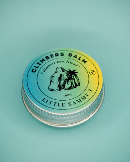 A 14ml tin of Little Sammy's Climbers Balm, closed and viewed from above, showcases the product's compact size. The top of the tin features a circular label with a gradient from blue to yellow, including an illustration of a boulder and a palm tree. The text reads 'CLIMBERS BALM - climbers best friend' with '14ml' and 'LITTLE SAMMY'S' prominently displayed, all against a soft blue background.