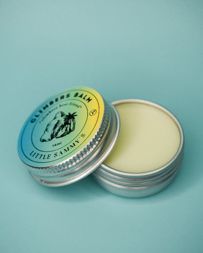 A 14ml tin of Little Sammy's Climbers Balm is presented open next to its lid, revealing the smooth, creamy balm inside. The lid, with its detailed label, rests beside the tin against a blue background, emphasising the product's petite size and the vibrant colours of the label.