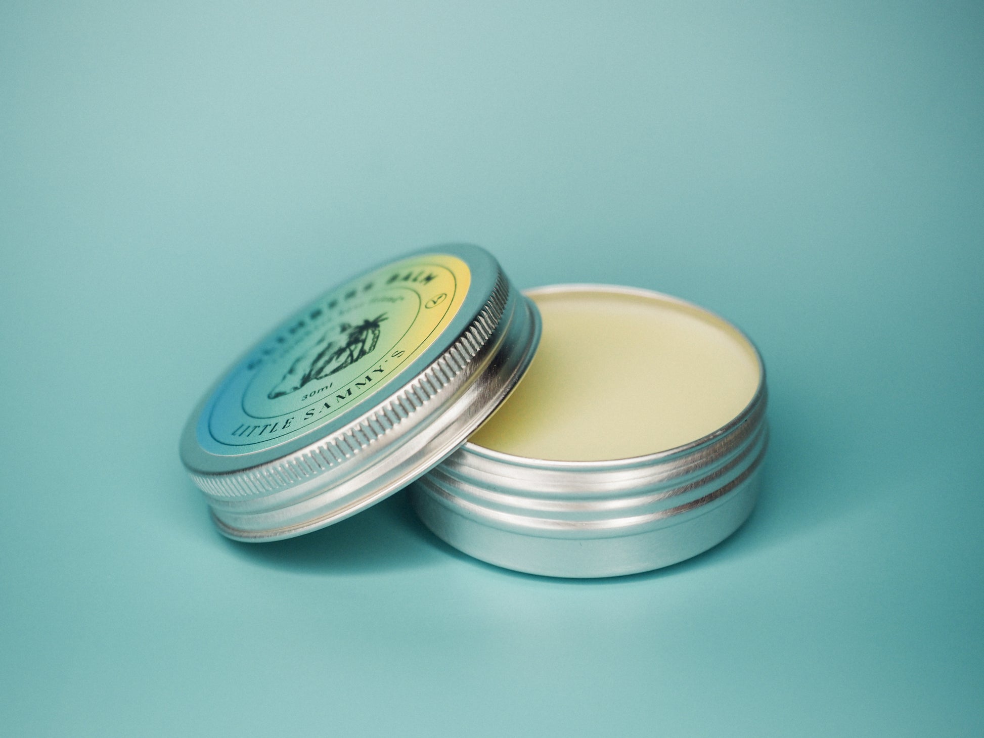 A scented 30ml tin of Little Sammy's Climbers Balm sits open to display its content against a soft blue background. The balm appears smooth and creamy. The tin lid, which is resting beside the base, features a vintage-style label with green and yellow accents and the image of a boulder and a palm tree, symbolising the brand.