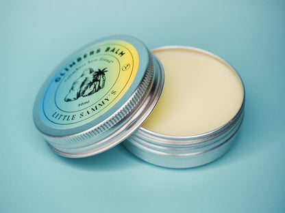 A 30ml tin of Little Sammy's Climbers Balm is shown open, with the lid angled to reveal the vibrant label. The label features a gradient of blue to yellow with a central logo depicting a boulder and a palm tree, symbolising a rock to be climbed and natural ingredients. The product's name is encircled around the logo, and the creamy texture of the balm is visible against the tin's reflective interior. All set against a tranquil blue background that complements the tin's colours.