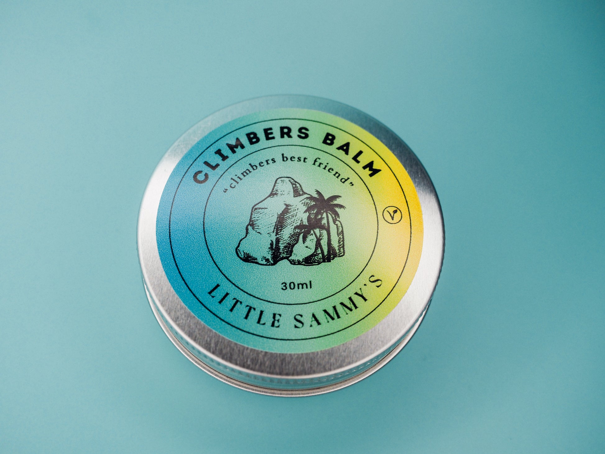 Close-up of a 30ml tin of Little Sammy's Climbers Balm, showcasing the lid's detailed label against a blue background. The label features a circular gradient of blue and yellow with the text 'CLIMBERS BALM - climbers best friend' encircling an illustration of a palm and a boulder. The product's name 'LITTLE SAMMY'S' is prominently displayed at the bottom, and a vegan symbol is also visible, indicating the balm's vegan-friendly nature.